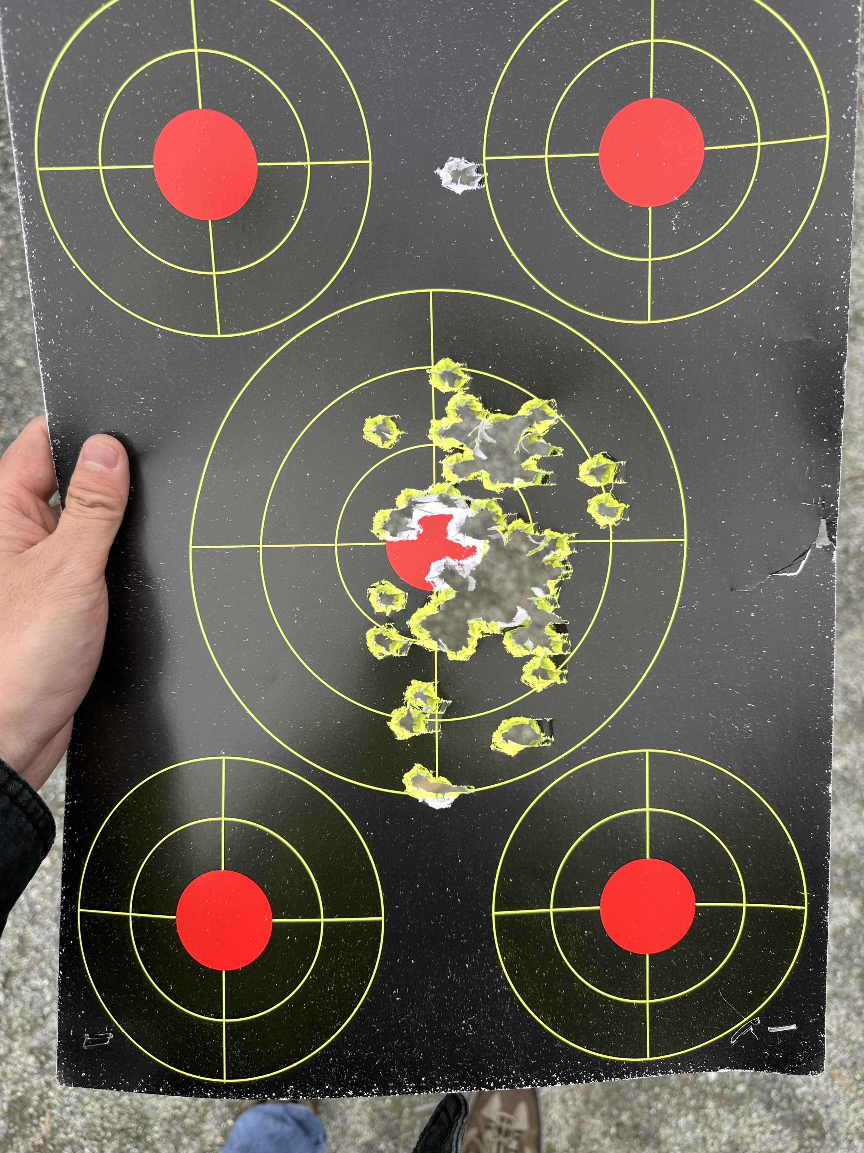 Five-target splatter shooting target held by a shooter, showing a heavily concentrated group of hits destroying the center bullseye with four outer targets untouched except for one stray hit.