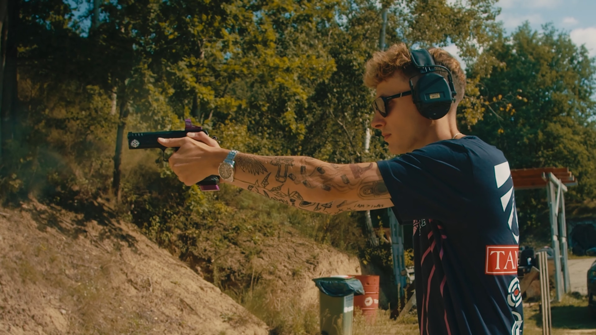 Young male shooter in ear protection and safety glasses firing a 1911-style pistol at an outdoor shooting range with a sand berm backstop.