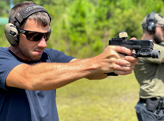Man in blue shirt shooting a pistol with red dot optic and weapon light at an outdoor range, another shooter visible in background.