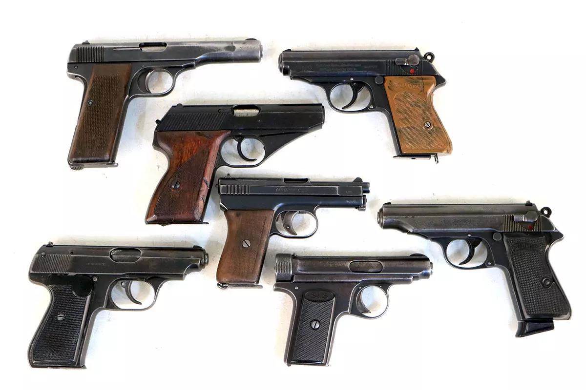 Six pre-war European pocket pistols laid out on a white surface, including Walther PP variants, a Mauser HSc, and several other blowback-operated compact pistols with wood and black synthetic grips.