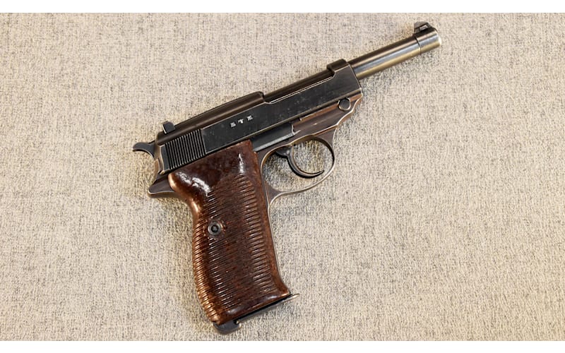 Blued semi-automatic pistol with checkered brown wood grips lying on a beige fabric surface, left side visible showing slide markings.