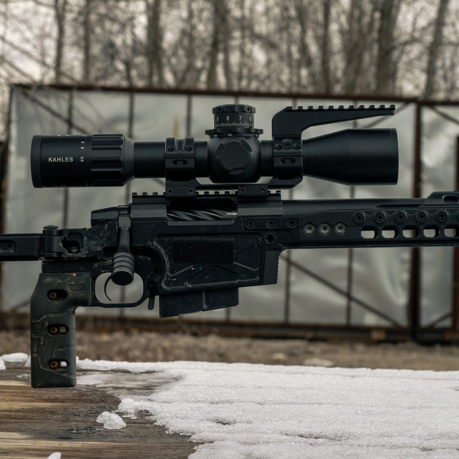 A Kahles riflescope mounted on a black bolt-action chassis rifle resting on a snow-dusted wooden surface outdoors.