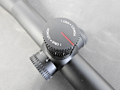 Close-up of a rifle scope turret with white text reading '1 click = 1/2 MOA' on a knurled black adjustment dial.