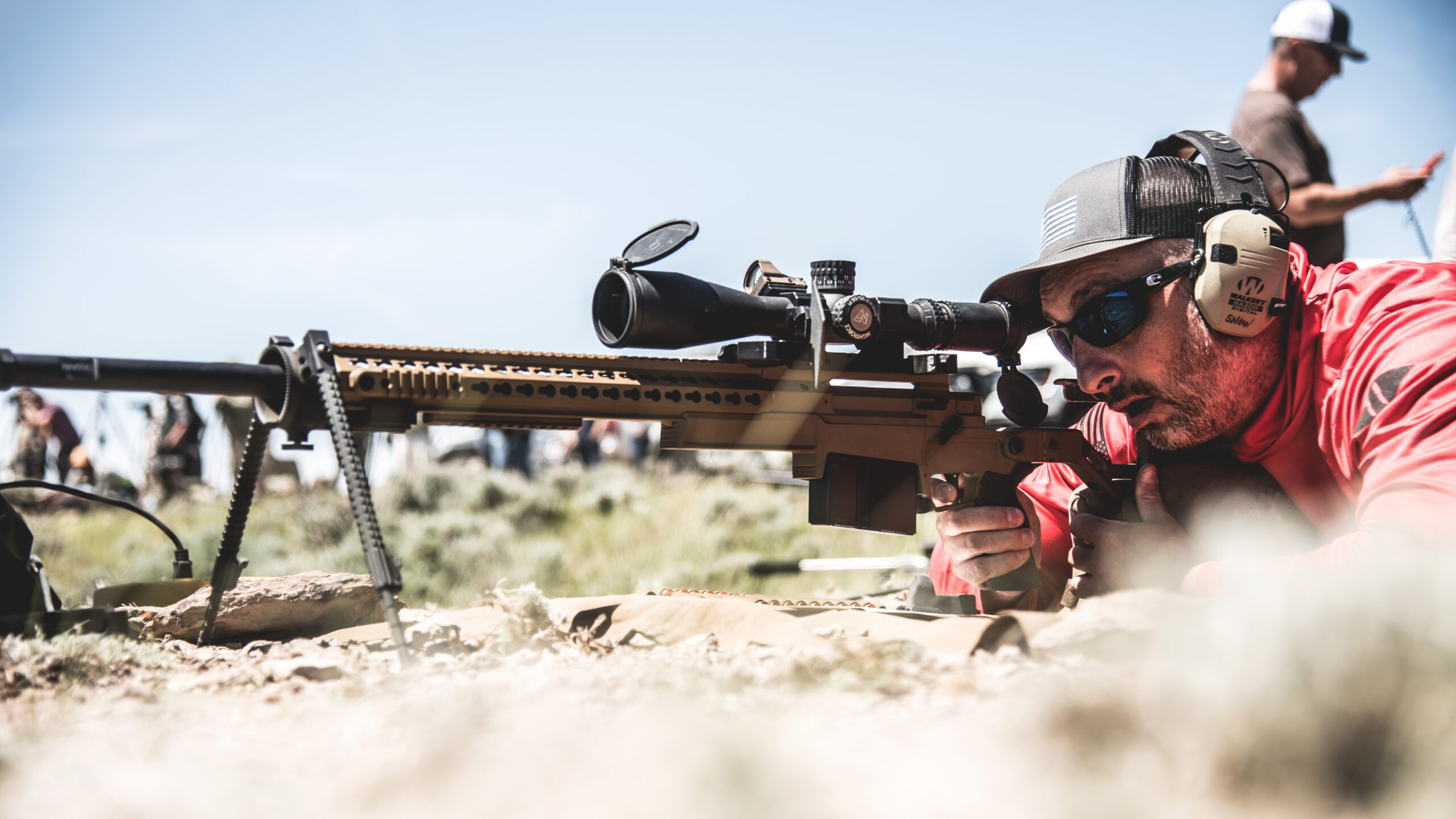 Shooter lying prone behind a tan bolt-action precision rifle with bipod and large scope at an outdoor long-range shooting event.