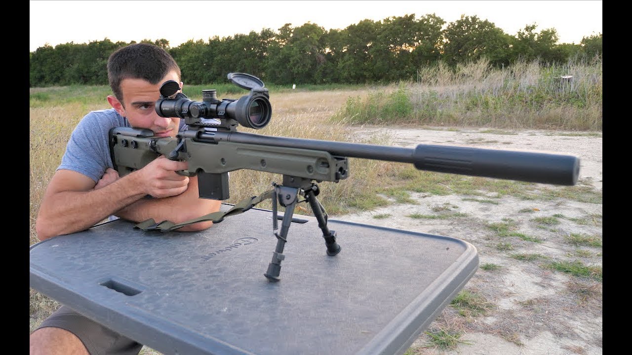 Shooter aiming a suppressed bolt-action rifle with a scope and bipod from a bench at an outdoor range.