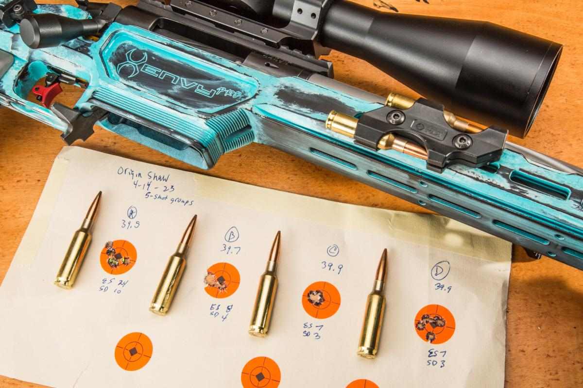 Bolt-action precision rifle in teal chassis resting above a handwritten target sheet showing four five-shot groups with chronograph data, alongside four rifle cartridges.