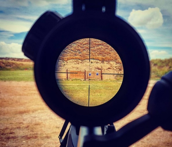 Shooter's eye view through a rifle scope with a duplex crosshair reticle, with paper targets visible downrange at an outdoor shooting range.