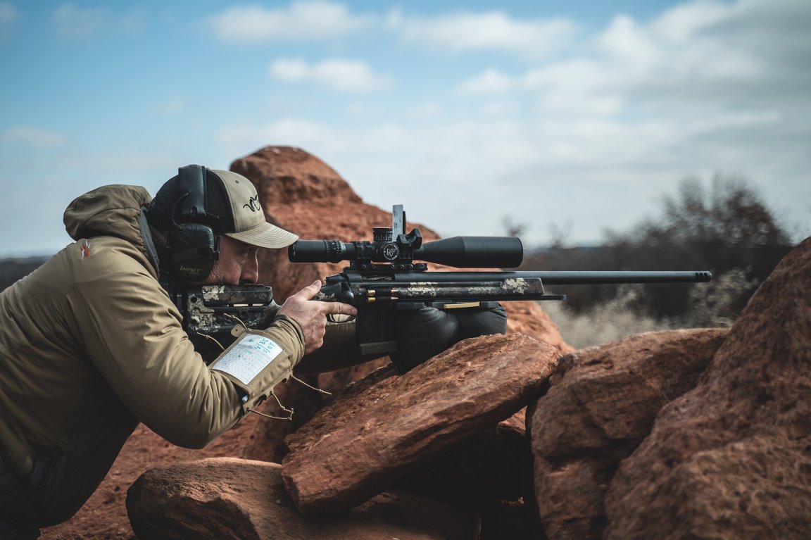 Male shooter prone over red rock barricade aiming a precision bolt-action rifle with a large scope, wearing electronic ear protection and a wrist-mounted dope card.