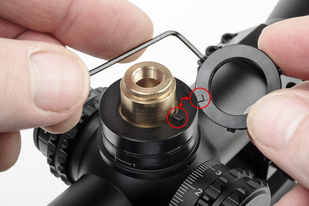 Close-up of hands using a tool to adjust a brass zero stop mechanism on a rifle scope turret, with red circles highlighting alignment marks.