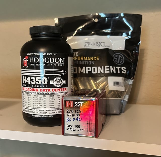 A bottle of Hodgdon H4350 rifle powder, a box of Hornady 130 grain SST .277 inch bullets, and a bag of SIG Sauer 270 Win brass cases arranged on a shelf.