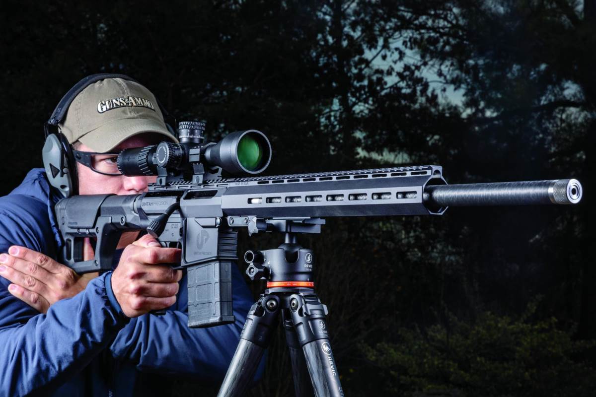 Shooter aiming a Uintah Precision AR-style precision rifle with a magnified scope, supported on a Revic tripod, outdoors at night.