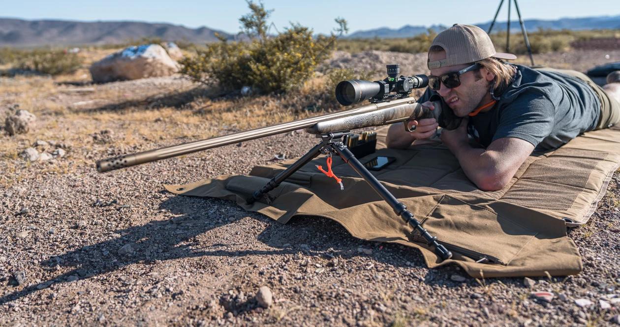 Shooter lying prone in a desert environment, aiming a long-range bolt-action rifle on a bipod with a large scope mounted on top.