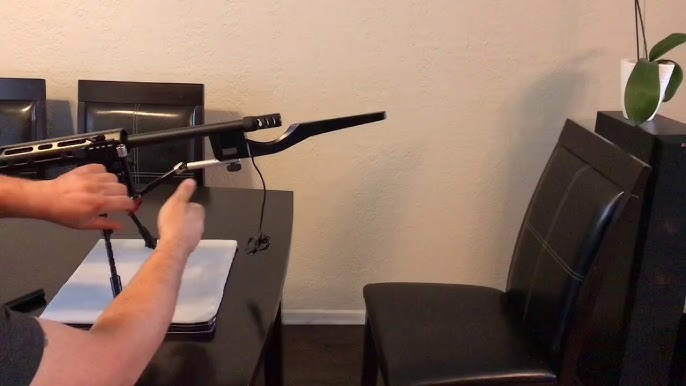 Hands holding a bolt-action rifle with a MagnetoSpeed chronograph sensor attached to the barrel over a table.