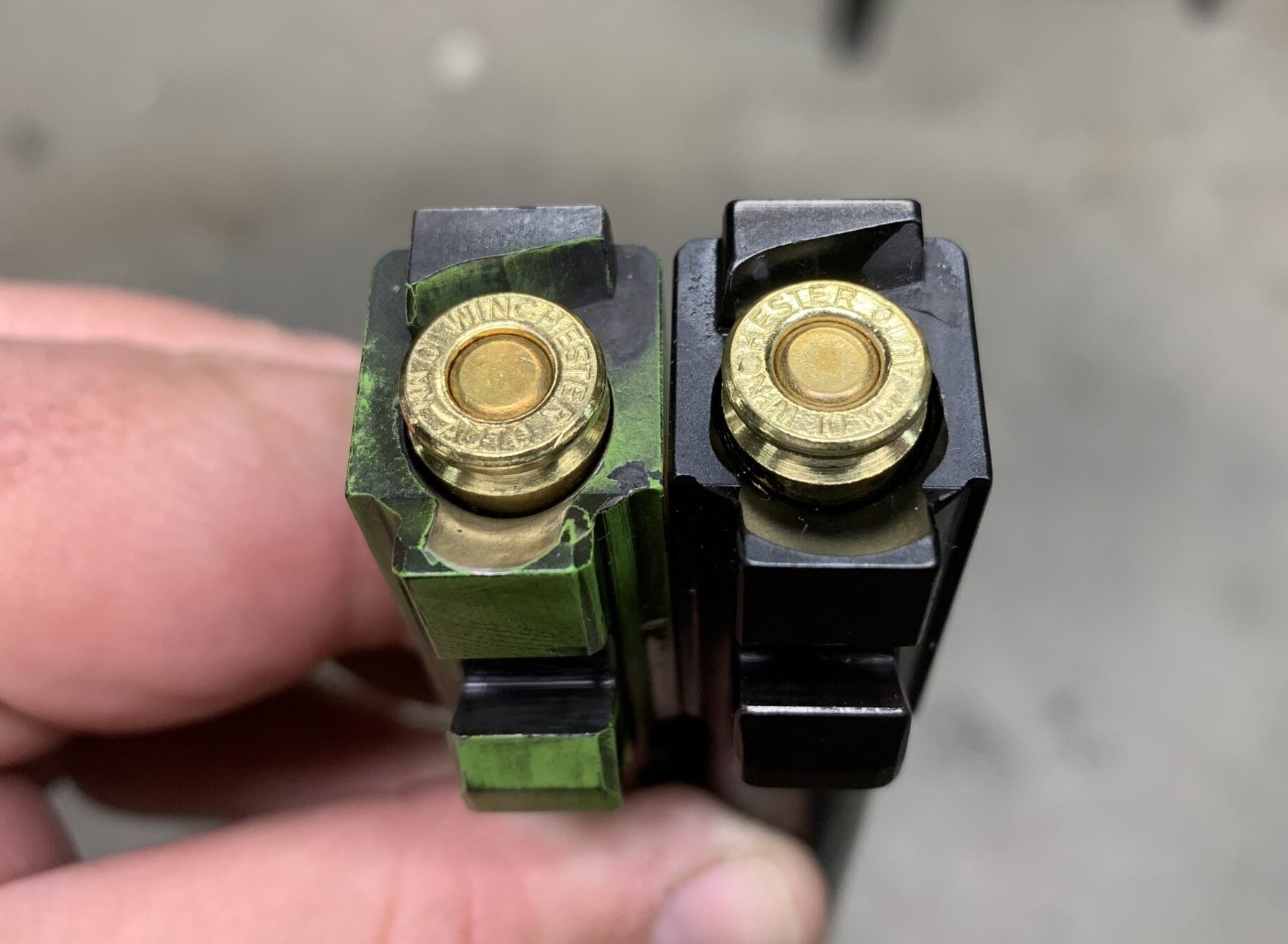Two pistol magazines side by side showing Winchester brass cartridge headstamps, one green-anodized and one black