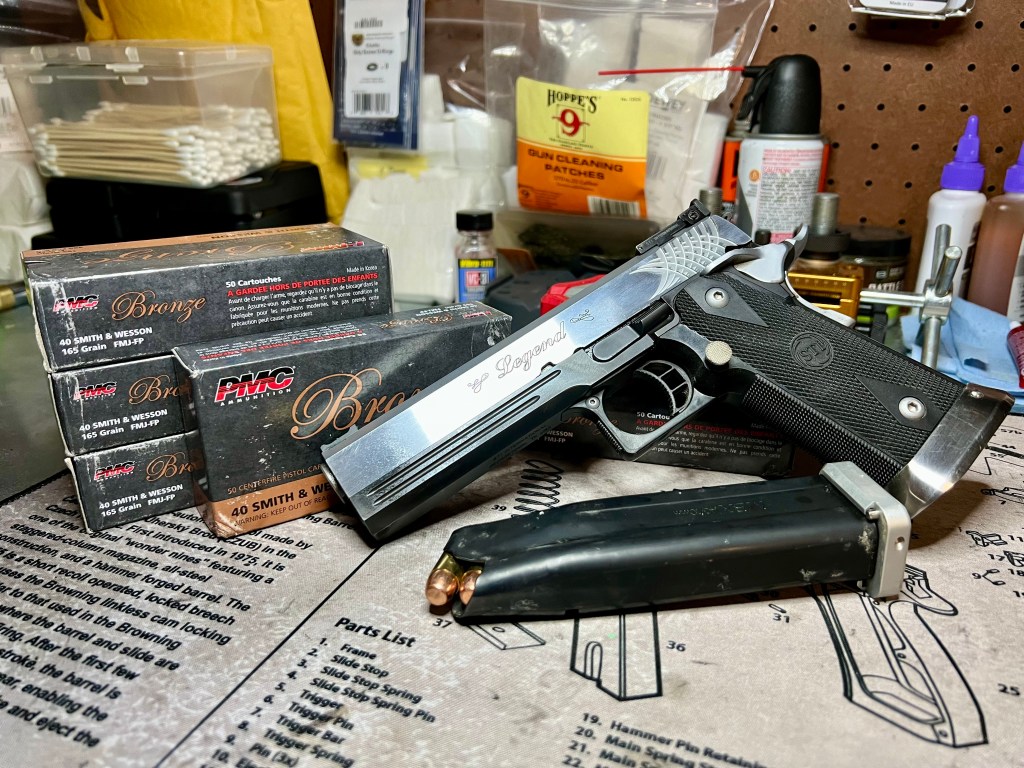 Stainless 2011 pistol with two loaded double-stack magazines on a workbench, next to boxes of PMC Bronze .40 S&W ammunition and Hoppe's cleaning supplies.