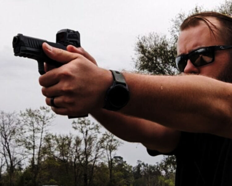 Shooter in sunglasses firing a red-dot-mounted semi-automatic pistol at full extension with a two-handed grip outdoors.