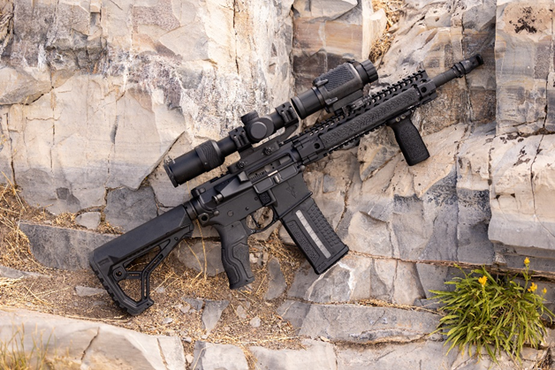 Black AR-15 style carbine with a clip-on thermal optic mounted in front of a day scope, resting against a rock outcropping outdoors.