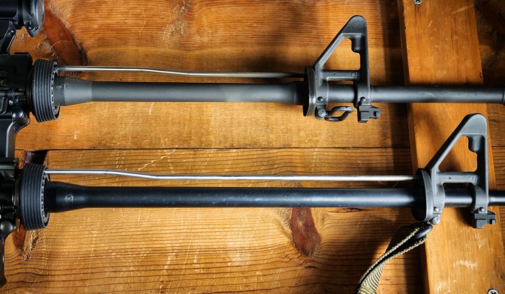 Two AR-15 barrels compared side by side on a wood surface, showing a thinner pencil-profile barrel above and a heavier-profile barrel below, both with A2-style front sight bases.