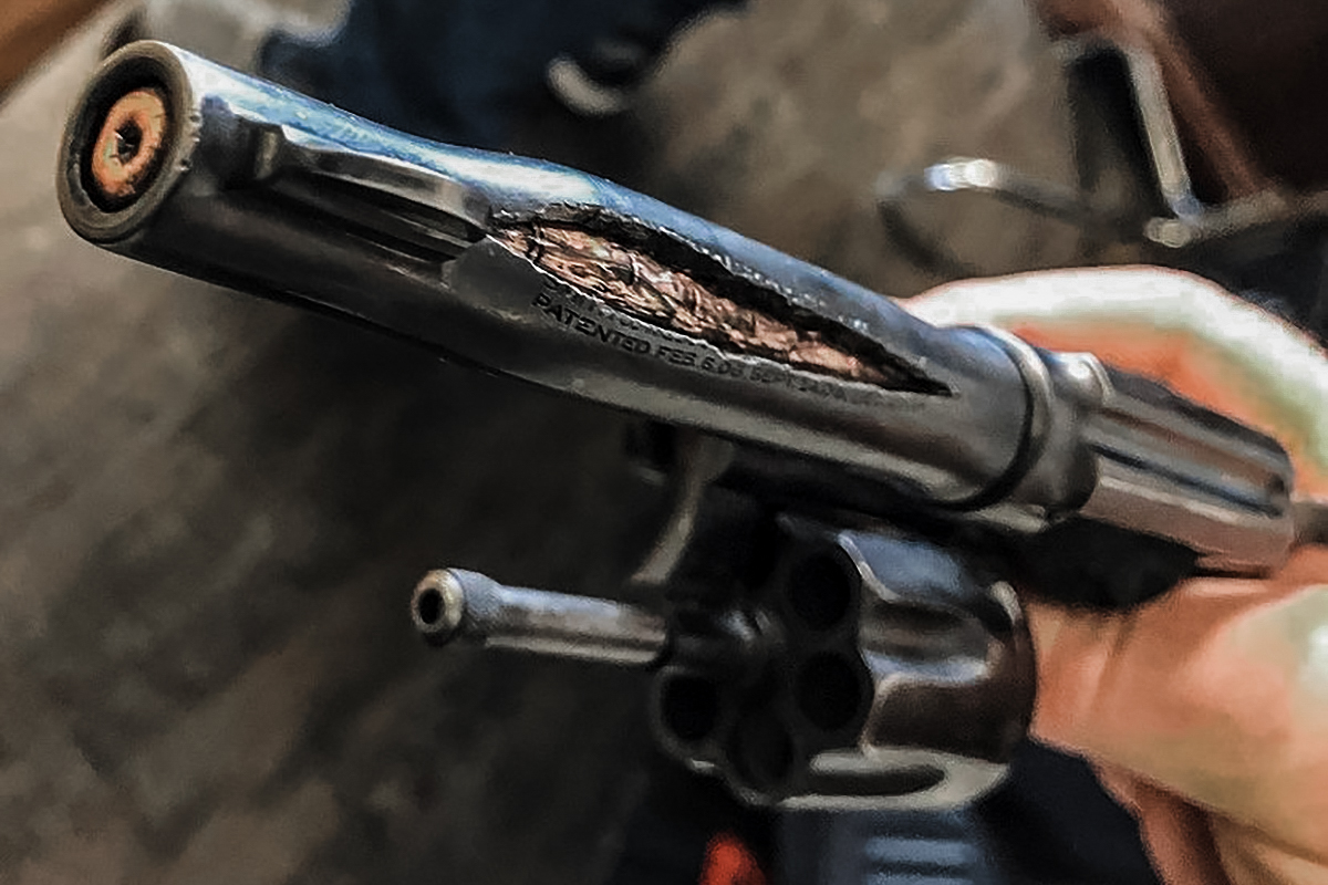 Close-up of a revolver barrel split open and torn apart by an internal pressure failure, with visible metal deformation along the top strap.