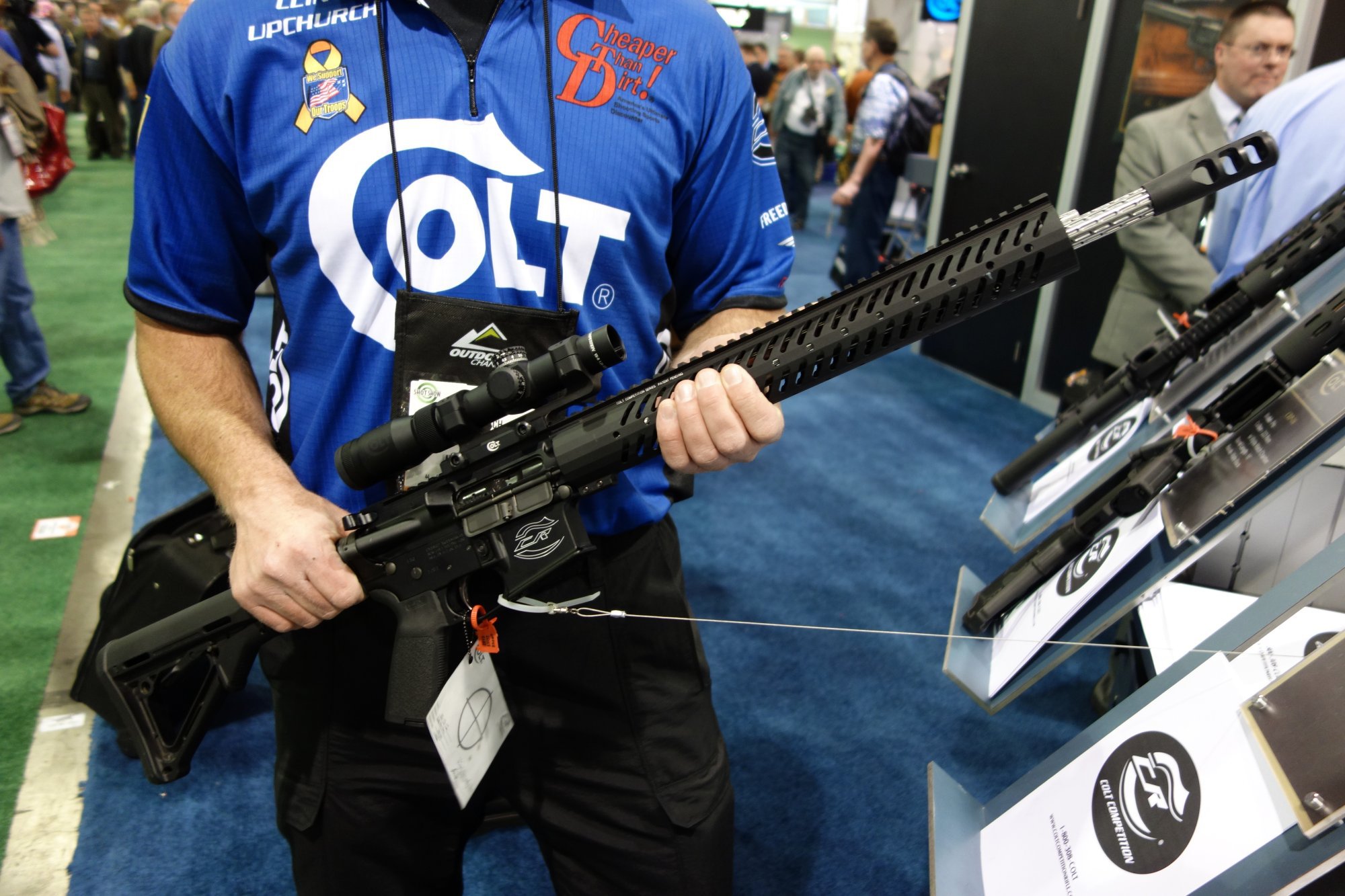 Person in Colt jersey holding a Colt Competition AR-15 with free-float rail and optic at a firearms trade show.
