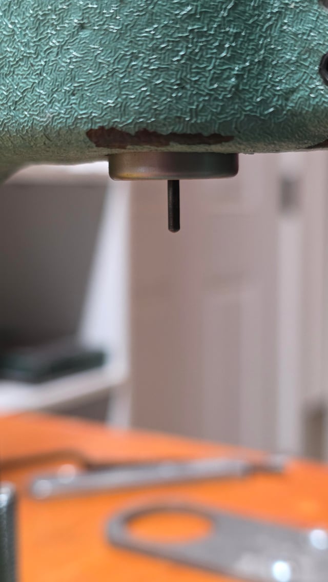 Close-up of a decapping pin protruding from the bottom of a reloading die secured in a green bench vise, with metal reloading parts on an orange surface below.