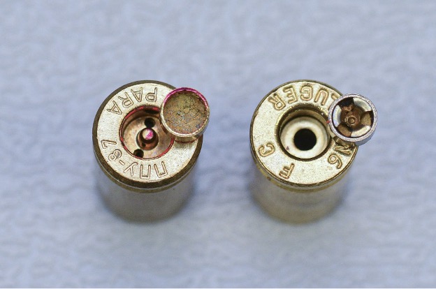 Bottom view of two spent brass cartridge cases side by side showing the difference between a Berdan-primed case with two flash holes and a Boxer-primed case with a single central flash hole.