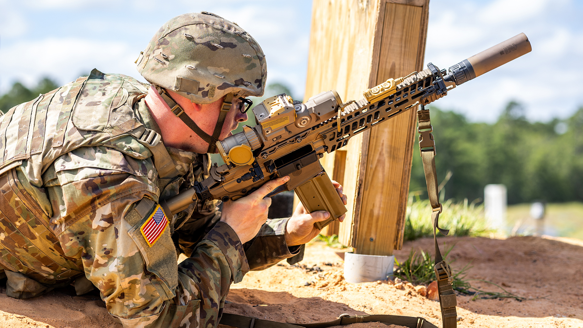 U.S. Army soldier in MultiCam uniform firing a suppressed XM7 rifle with XM157 optic from a prone position at an outdoor range.