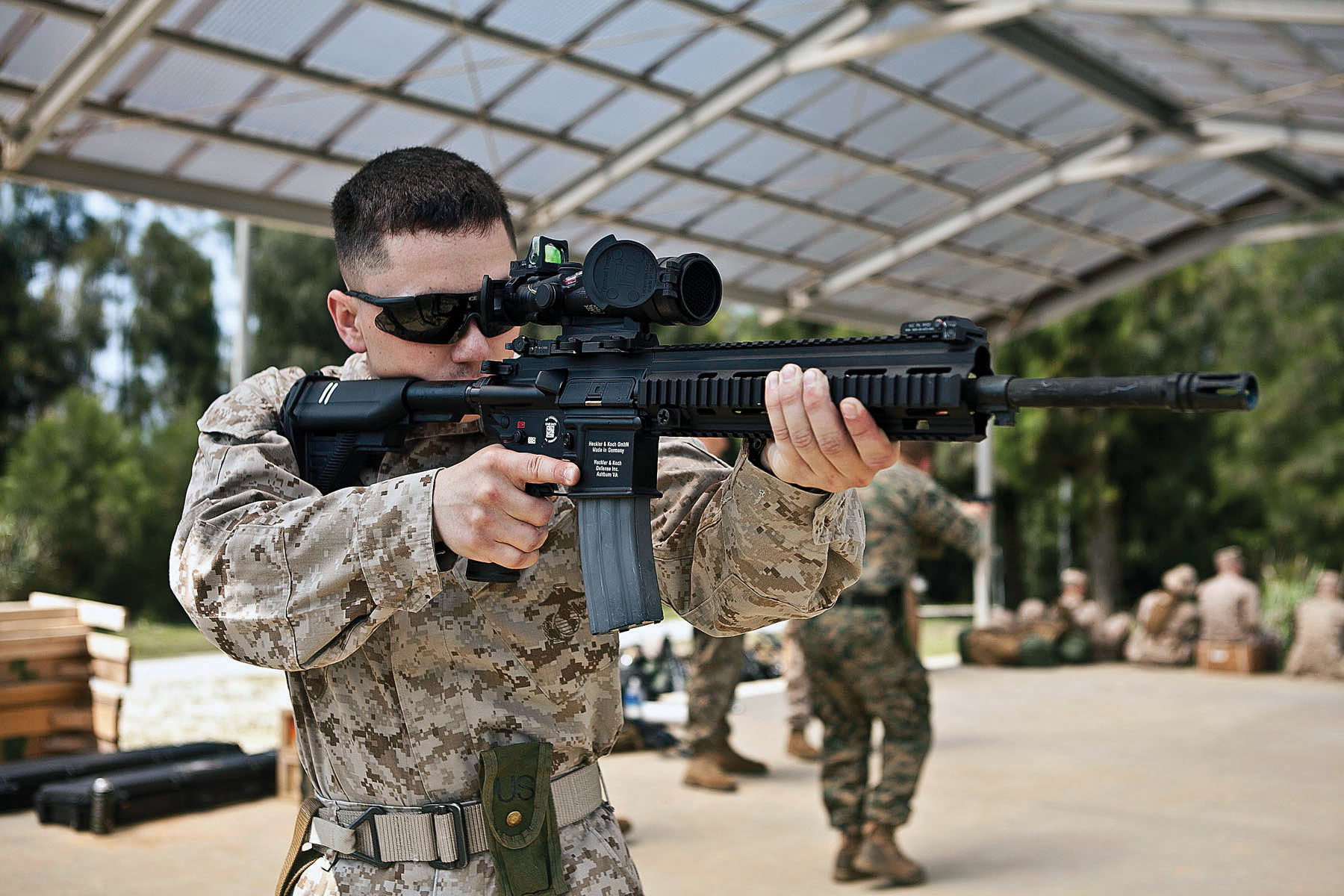 U.S. Marine aiming an HK416-pattern rifle with optics at an outdoor covered range, other Marines visible in the background.