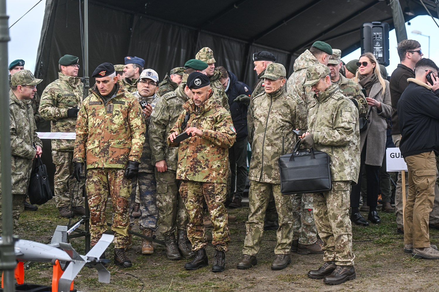 Group of multinational military officers in various camouflage uniforms gathered under a field tent at an outdoor military exercise.