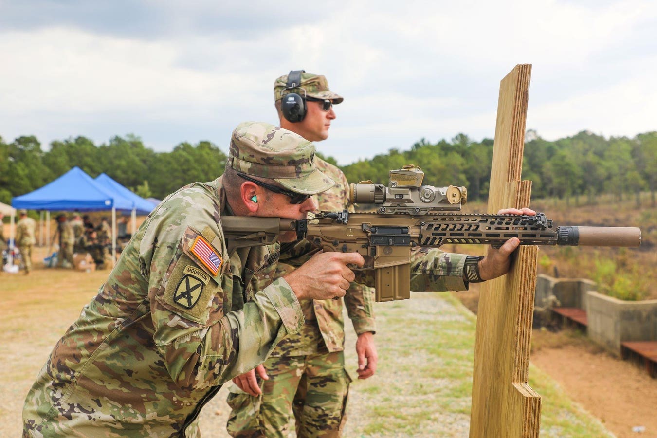 U.S. Army soldier from 10th Mountain Division aiming an XM7 rifle with suppressor and optic from a barricade position at an outdoor range.