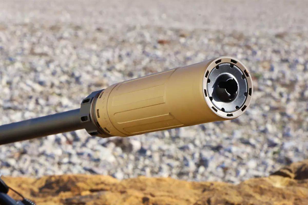 Close-up of a flat dark earth suppressor mounted on a black rifle barrel, viewed from the muzzle end at an outdoor range.