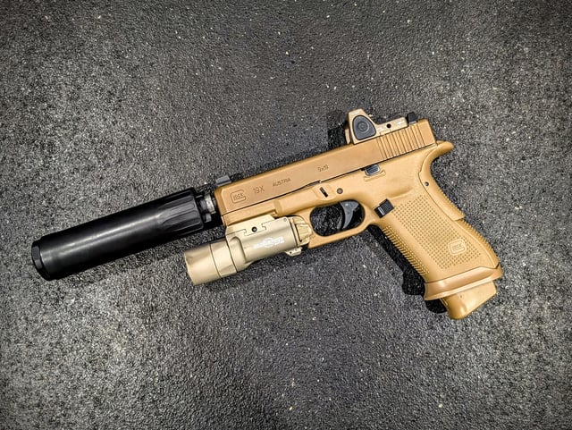 FDE Glock 19X pistol with suppressor, weapon light, and red dot sight laid flat on a dark surface.