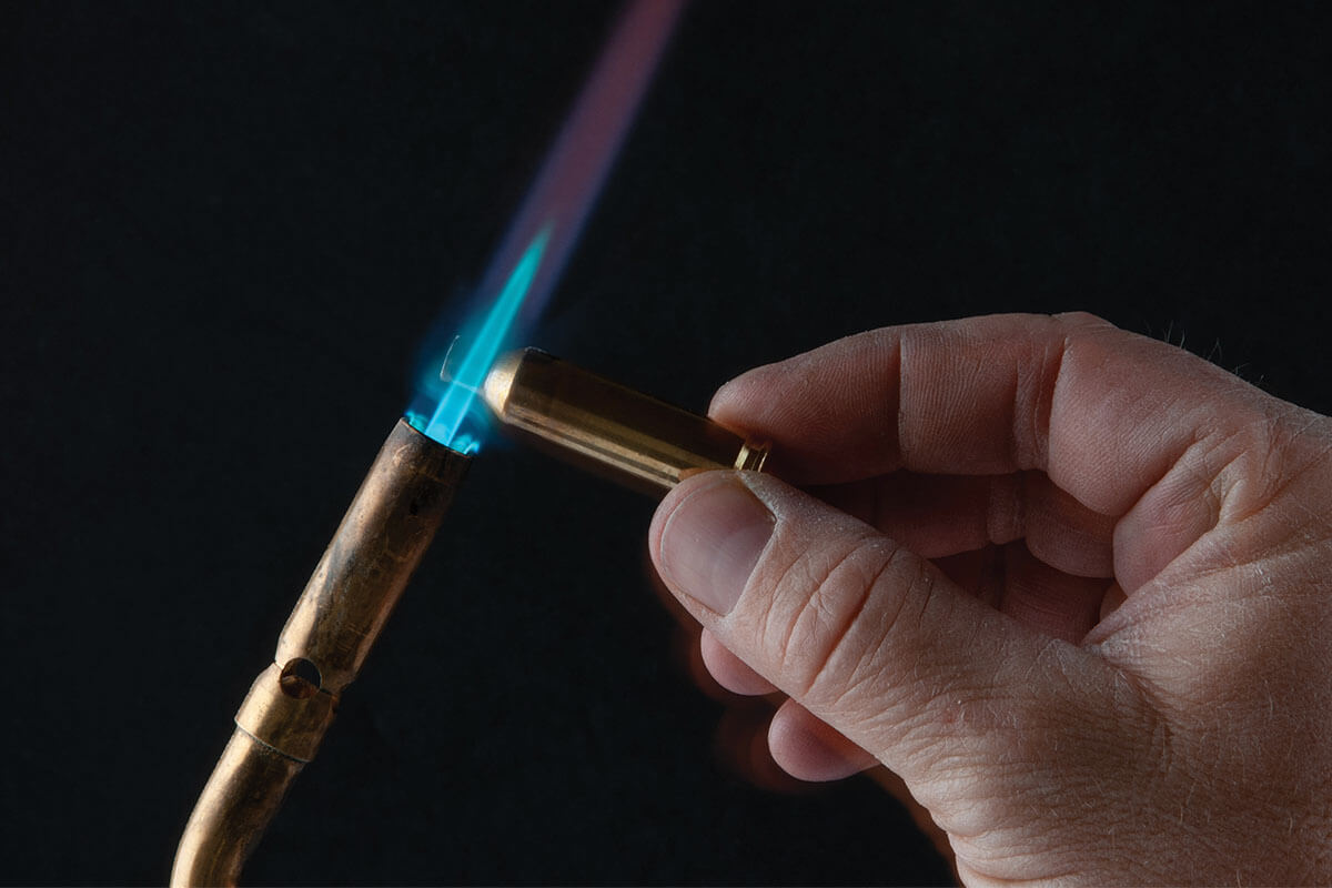 Hand holding a brass cartridge case by the head while a torch flame heats the neck and shoulder for annealing.