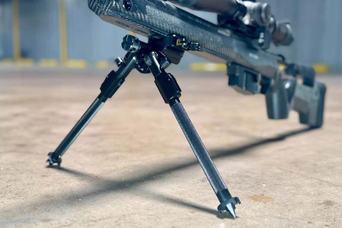 Close-up of an Atlas bipod mounted on a bolt-action rifle in an MDT chassis, legs extended and resting on a concrete surface.