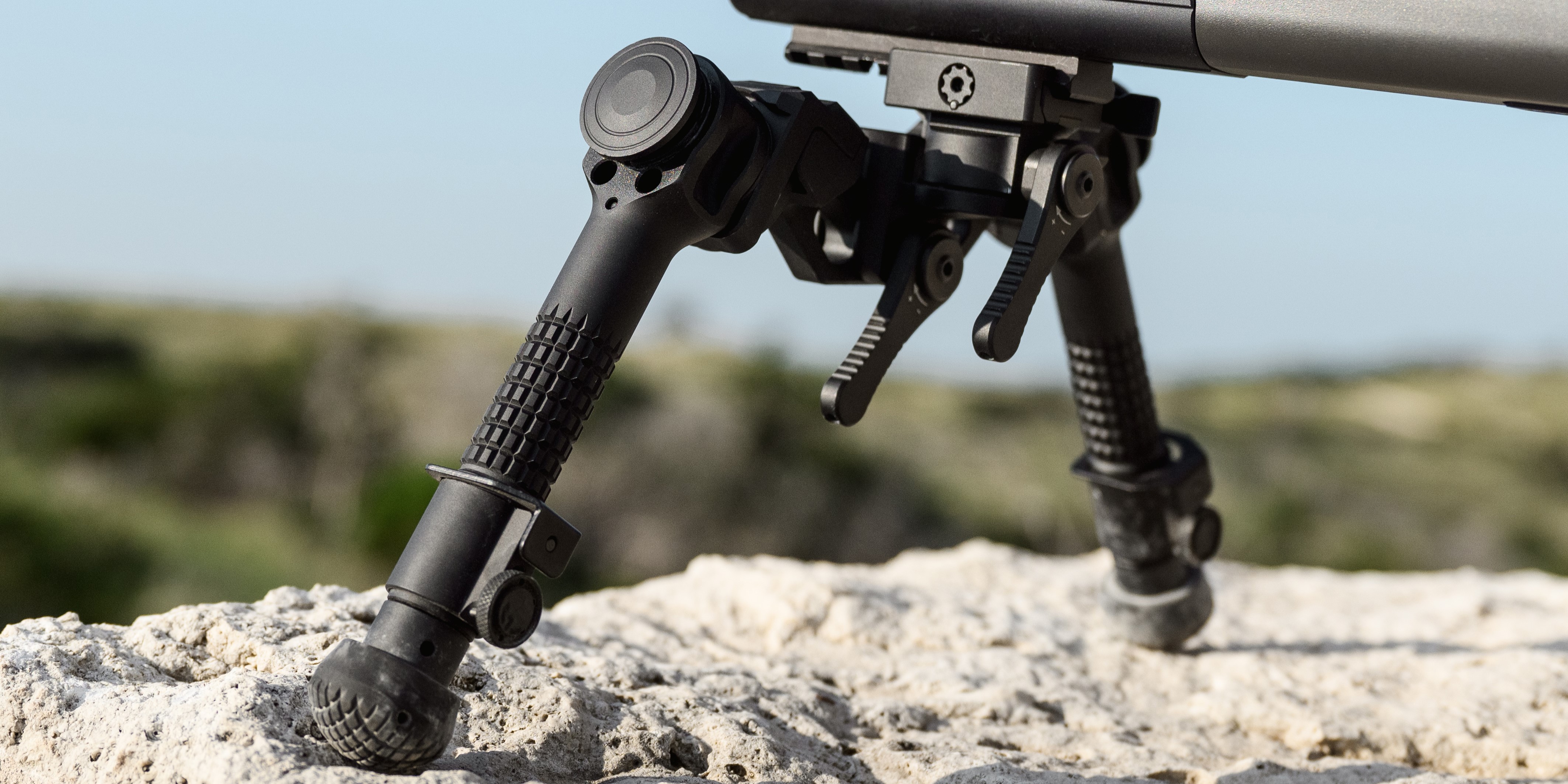 Close-up of a black tactical bipod with knurled adjustable legs and rubber feet mounted to a precision rifle, resting on a limestone rock surface outdoors.