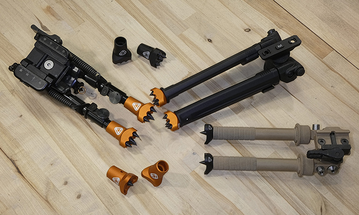 Two rifle bipods displayed on a wood surface alongside multiple interchangeable foot attachments including spiked orange feet and rubber feet options.