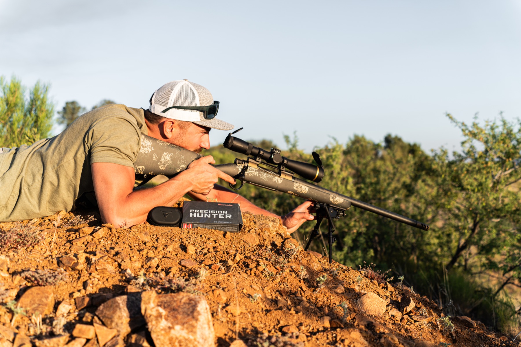 Hunter shooting prone on a rocky hillside with a camo bolt-action rifle on a bipod, Hornady Precision Hunter ammo box in the foreground.