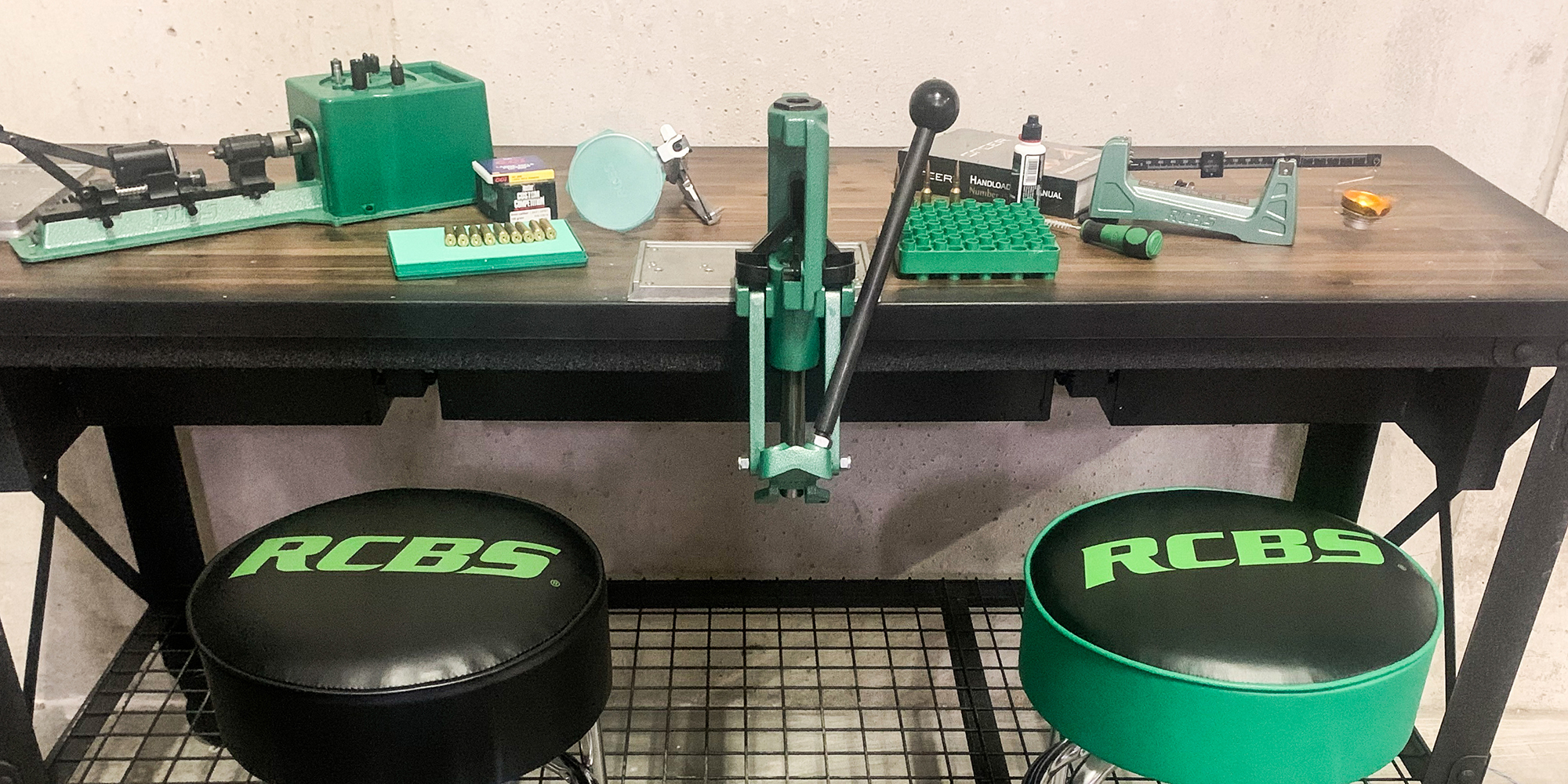 RCBS reloading bench setup with a single-stage press, case trimmer, beam scale, loading blocks, and reloading manual on a wood-top workbench, with two RCBS branded stools in front.