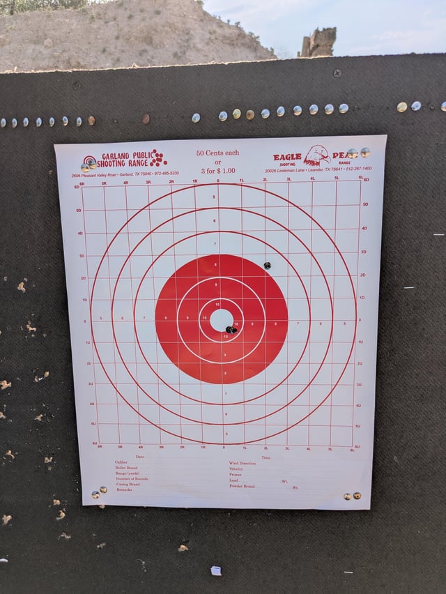 Paper bullseye target with two bullet holes, one near center and one slightly high, shot at an outdoor range.