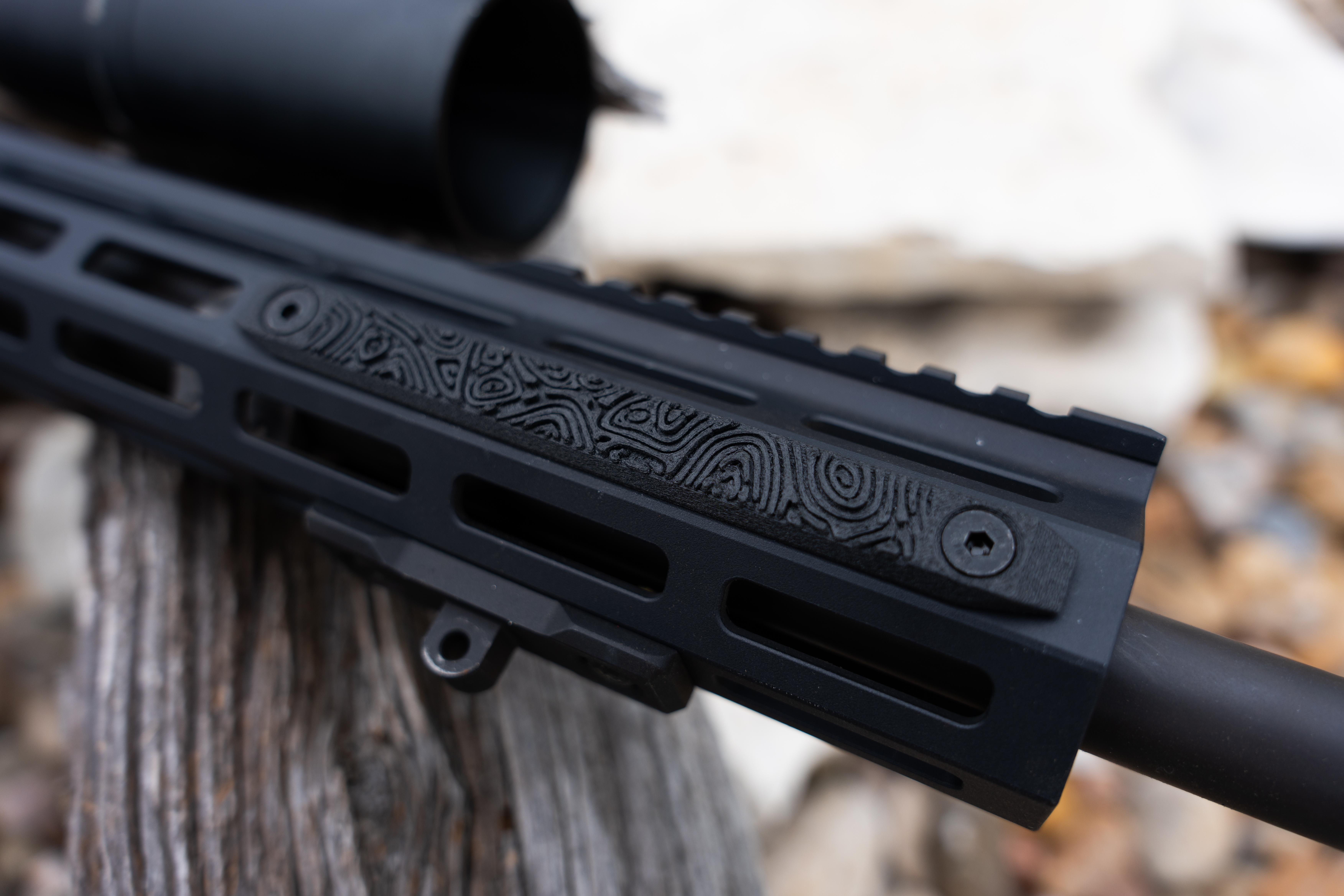 Close-up of a black AR-15 M-LOK handguard with a decorative topographic-patterned rail panel insert, with a suppressor and scope visible in the background.