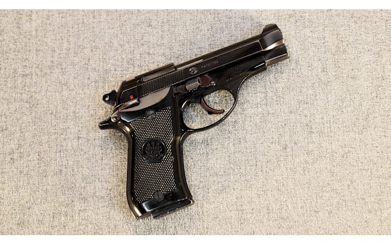 Blued Beretta 81 or 84 series compact pistol with checkered grips, shown from the left side on gray fabric