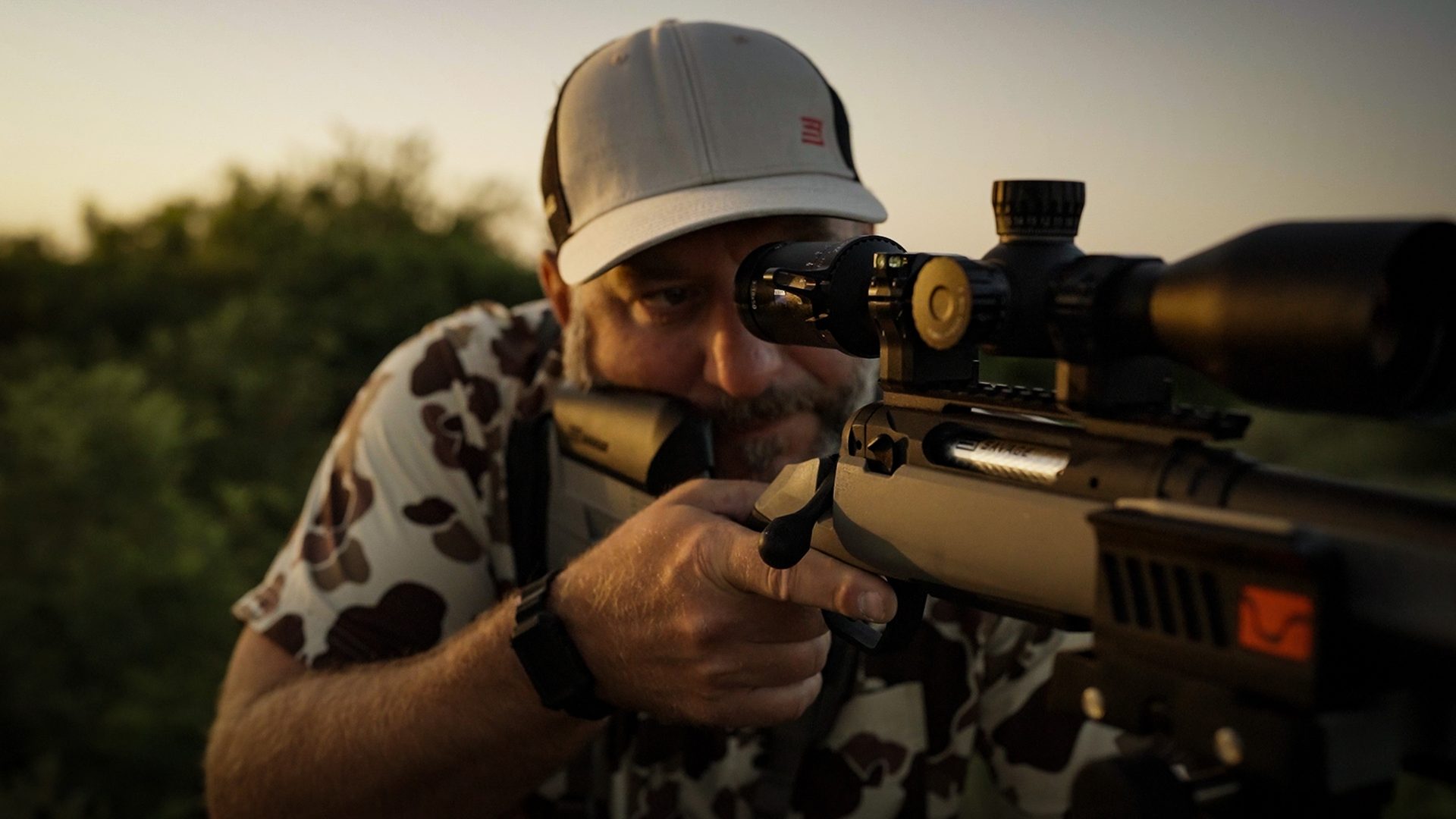 Man in camo shirt aiming a Savage 110 bolt-action rifle with a tan stock and riflescope outdoors at dusk.