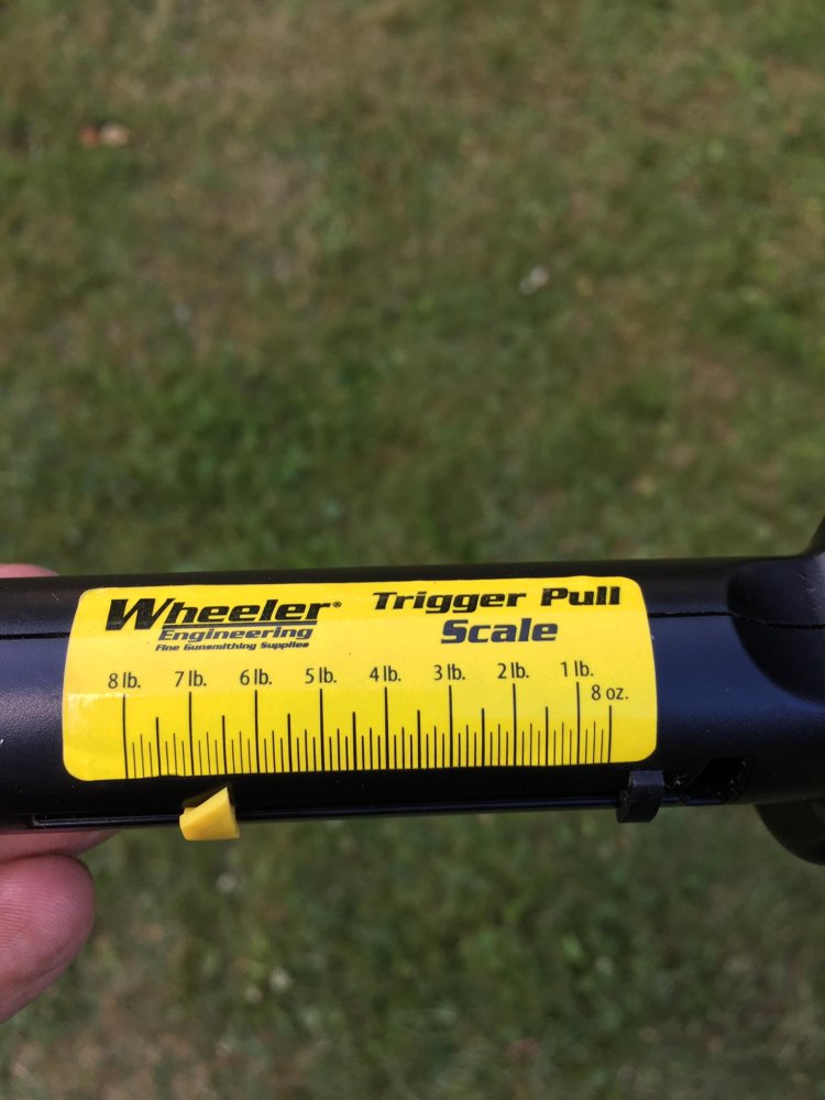 Hand holding a Wheeler Engineering analog trigger pull scale with a yellow label showing measurements from 8 ounces to 8 pounds.
