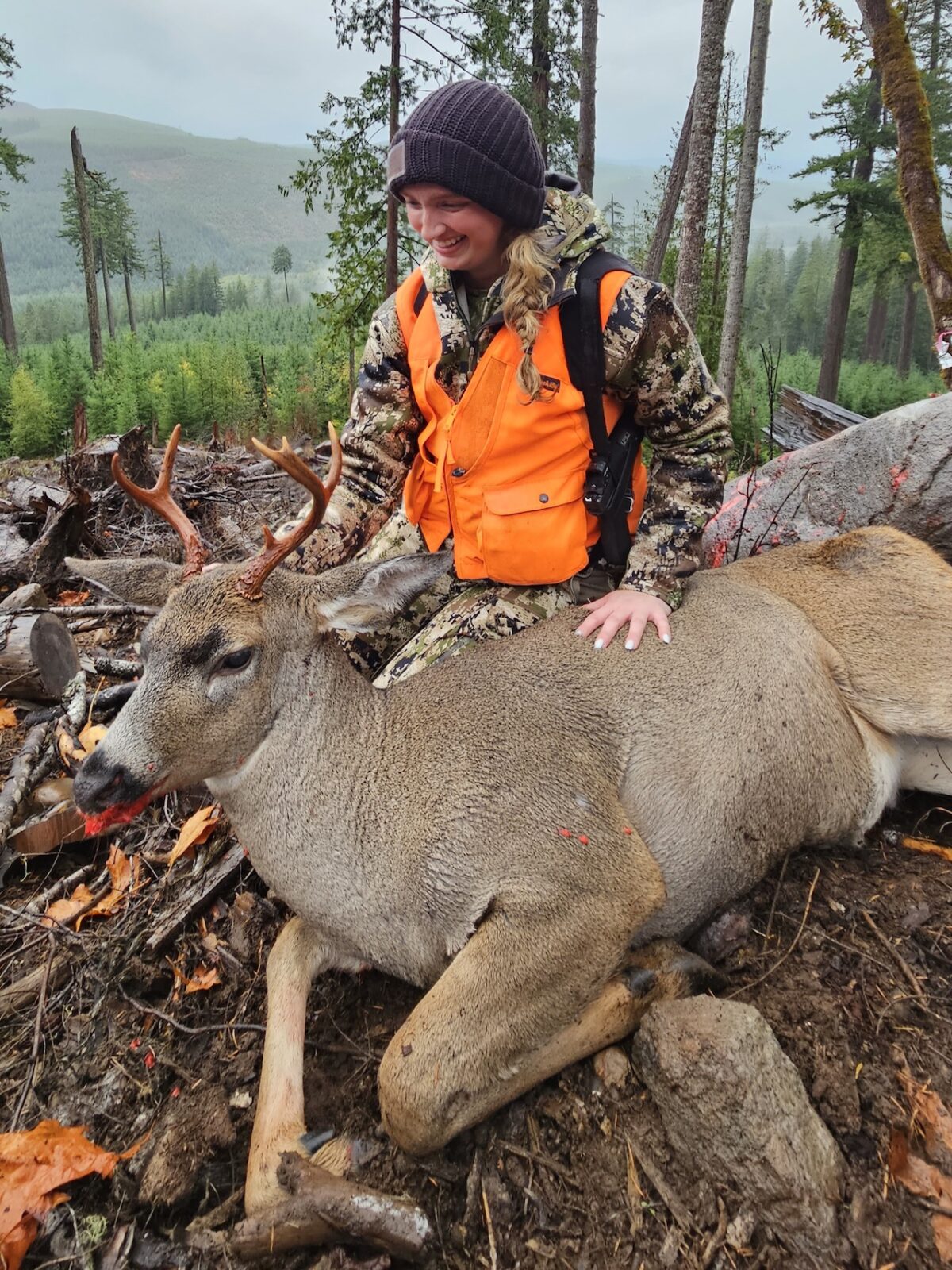 Young female hunter in blaze orange vest and camo kneeling behind a harvested whitetail buck in a forested Pacific Northwest clearcut.