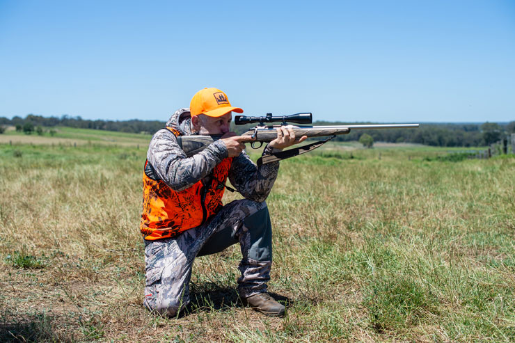 Hunter in blaze orange vest kneeling in a grassy field, aiming a stainless bolt-action rifle with a scope toward the horizon.