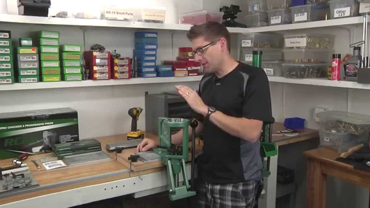 Man standing at a reloading bench demonstrating an RCBS Rock Chucker single-stage press, with shelves of reloading components and die boxes in the background.