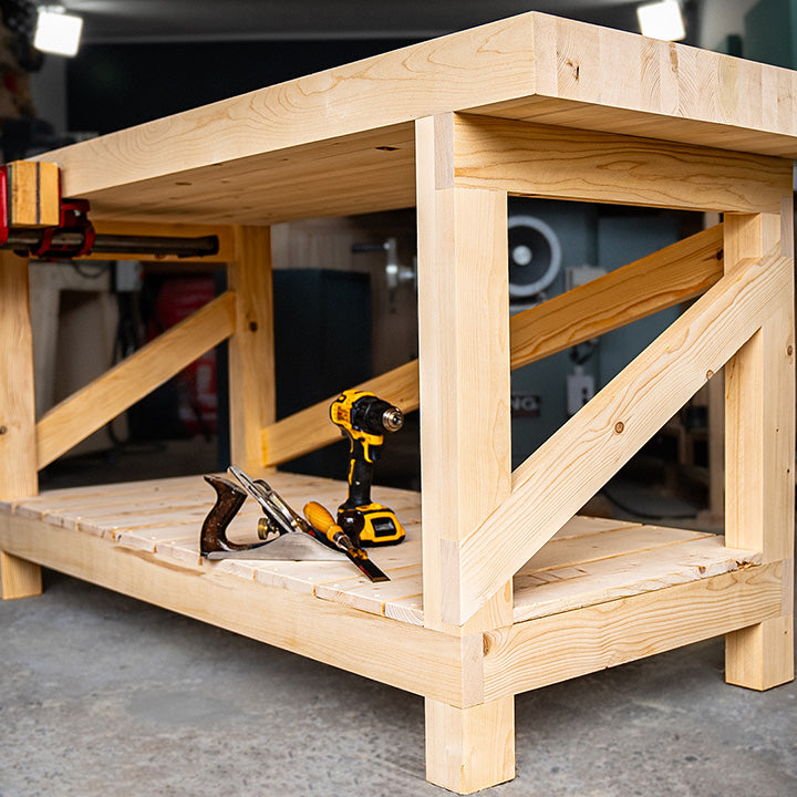 Sturdy wooden workbench with X-pattern diagonal cross-bracing on the side panels, lower shelf, and a cordless drill and hand plane stored underneath.