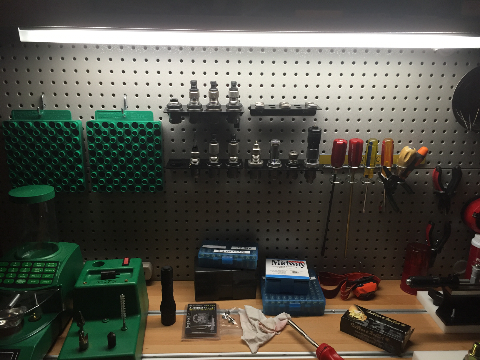 Reloading bench with pegboard wall organizer holding reloading dies, case trays, screwdrivers, and pliers above a workbench with an RCBS press, ammo trays, and Sellier & Bellot ammunition box.
