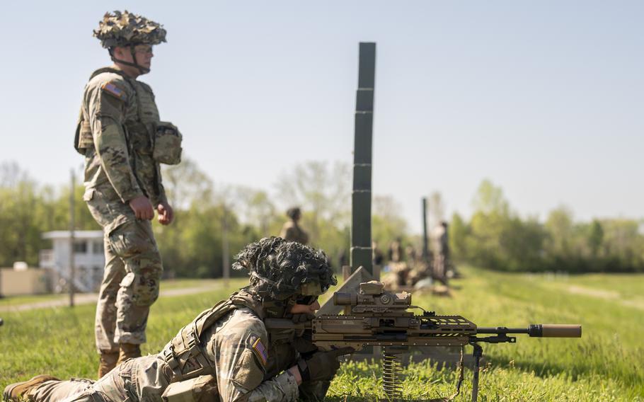 U.S. Army soldier in prone position firing an XM250 automatic rifle during a field training exercise while another soldier stands watch.