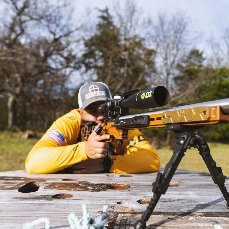 Shooter in yellow shirt aiming a gold chassis bolt-action rimfire rifle on a bipod from a wooden bench outdoors.